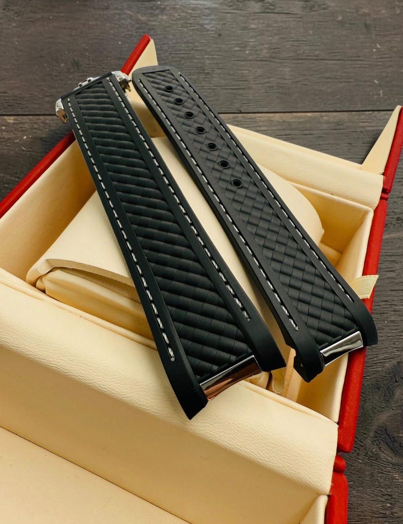 May include: Two black watch straps with white stitching and silver-tone hardware are displayed inside a red and cream-colored box. The straps have a textured pattern and are made of a flexible material.