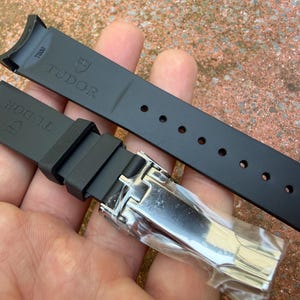 K&ouml;nnte beinhalten: Schwarzes Kautschuk-Uhrenarmband mit dem Markennamen "TUDOR" und Logo. Das Armband hat eine Edelstahlschlie&szlig;e und mehrere Verstelll&ouml;cher. Es ist f&uuml;r ein bestimmtes Uhrenmodell konzipiert.
