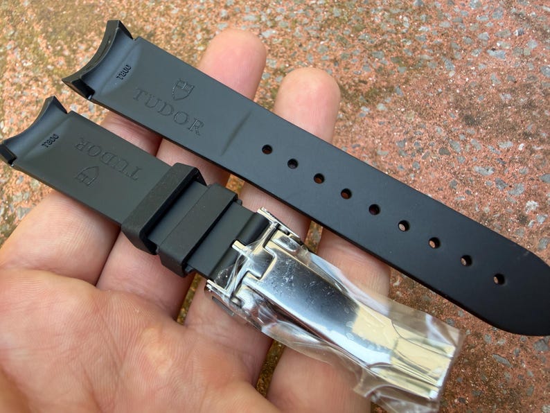 K&ouml;nnte beinhalten: Schwarzes Kautschuk-Uhrenarmband mit dem Markennamen "TUDOR" und Logo. Das Armband hat eine Edelstahlschlie&szlig;e und mehrere Verstelll&ouml;cher. Es ist f&uuml;r ein bestimmtes Uhrenmodell konzipiert.