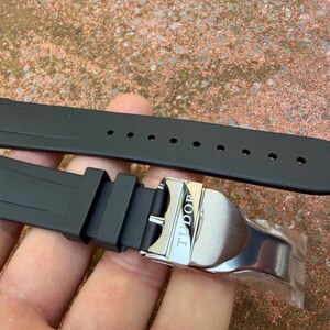 May include: A black rubber watch strap featuring a silver-toned metal deployment clasp with the word TUDOR engraved on the surface. The strap includes a series of circular adjustment holes and two rubber keepers to secure the excess band length.