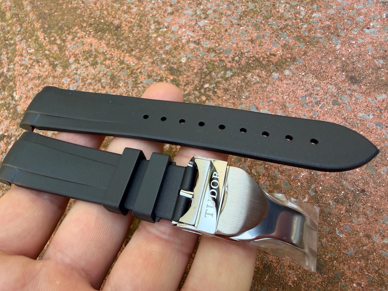 May include: A black rubber watch strap featuring a silver-toned metal deployment clasp with the word TUDOR engraved on the surface. The strap includes a series of circular adjustment holes and two rubber keepers to secure the excess band length.