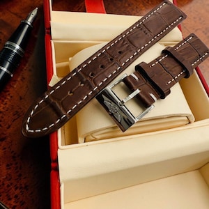 May include: A brown leather watch strap with a crocodile pattern and white stitching. The strap is displayed in a red and cream-colored box with a silver buckle.