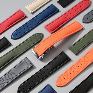 May include: A collection of colorful watch straps in various shades, including red, blue, green, and orange. The straps are made of a textured material and feature contrasting stitching. Some straps have a buckle, while others have a pin closure. The straps are arranged on a light gray surface.