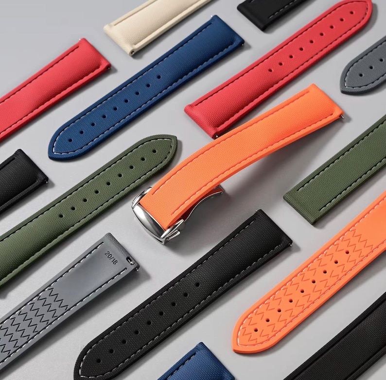 May include: A collection of colorful watch straps in various shades, including red, blue, green, and orange. The straps are made of a textured material and feature contrasting stitching. Some straps have a buckle, while others have a pin closure. The straps are arranged on a light gray surface.