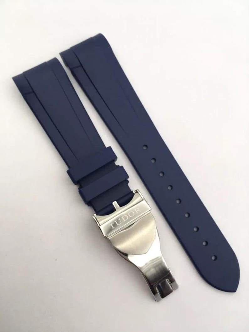 May include: A dark blue rubber watch strap featuring a silver-toned metal deployment clasp with the word TUDOR engraved on the surface. The strap consists of two separate pieces, one with a series of adjustment holes and the other with the clasp.