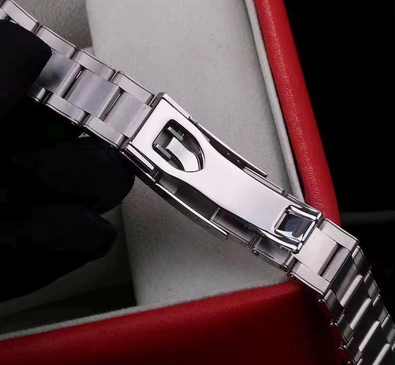 Pu&ograve; includere: Un cinturino per orologio in metallo color argento con finitura lucida e un meccanismo di chiusura deployante sicuro. Le maglie sono rettangolari e collegate in un design standard a tre pezzi, con una superficie liscia e riflettente.