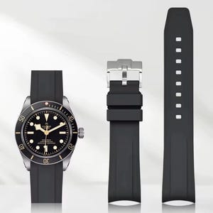 May include: A black wristwatch with a silver bezel and a black rubber strap. The watch face is black with white and gold accents. The image also shows a black rubber watch strap and a silver buckle.