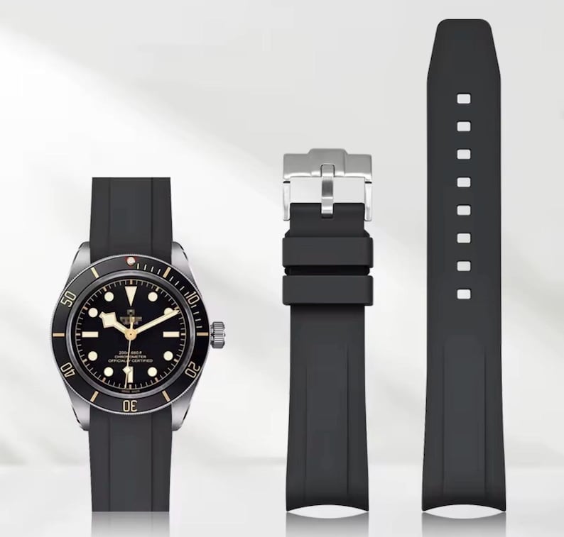 May include: A black wristwatch with a silver bezel and a black rubber strap. The watch face is black with white and gold accents. The image also shows a black rubber watch strap and a silver buckle.