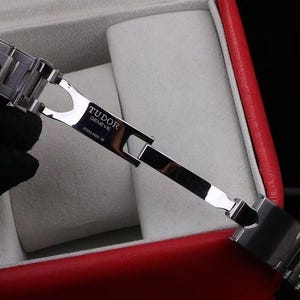 Pu&ograve; includere: Una chiusura per bracciale di orologio in acciaio inossidabile color argento in posizione aperta. La superficie metallica presenta il testo inciso TUDOR GENEVE e STEELINOX XR. Il meccanismo di chiusura &egrave; articolato con maglie lucide e design pieghevole.