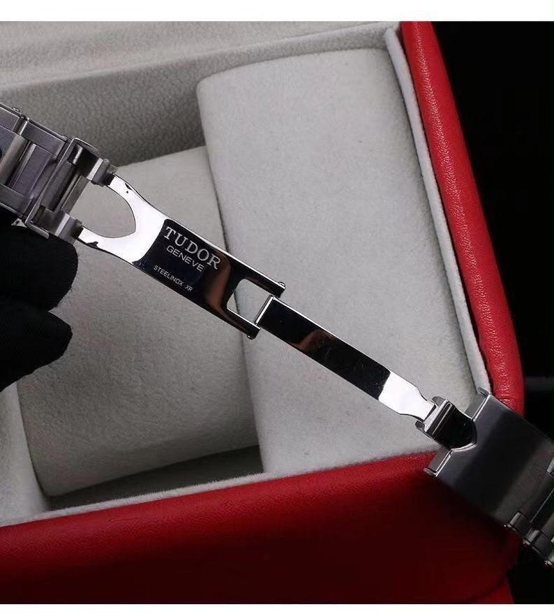 Pu&ograve; includere: Una chiusura per bracciale di orologio in acciaio inossidabile color argento in posizione aperta. La superficie metallica presenta il testo inciso TUDOR GENEVE e STEELINOX XR. Il meccanismo di chiusura &egrave; articolato con maglie lucide e design pieghevole.
