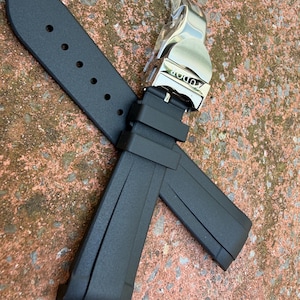 May include: A black rubber watch strap featuring a silver-toned metal deployment clasp with the Tudor brand name engraved on the surface. The strap includes a series of adjustment holes and two integrated rubber keepers for securing the band.