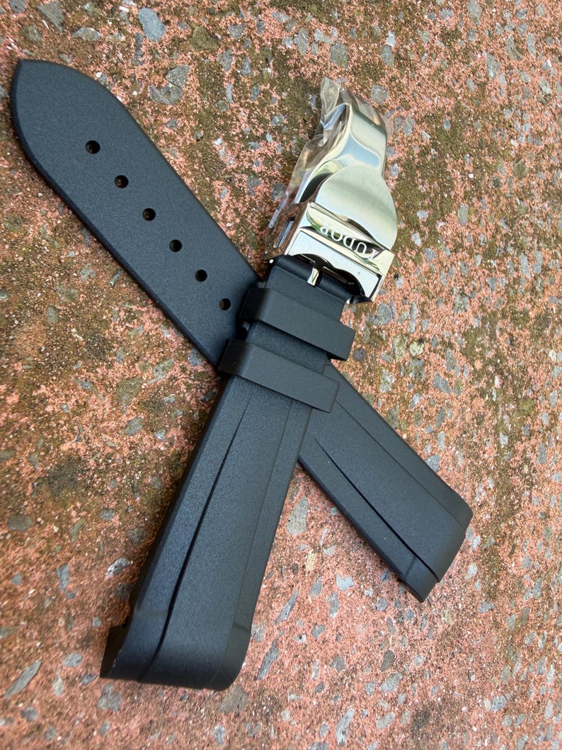 May include: A black rubber watch strap featuring a silver-toned metal deployment clasp with the Tudor brand name engraved on the surface. The strap includes a series of adjustment holes and two integrated rubber keepers for securing the band.