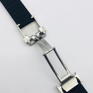 May include: A black rubber watch strap featuring a silver-toned stainless steel deployment clasp. The strap has a textured surface and the metal mechanism is shown in an open, extended position, revealing the hinge and locking components.