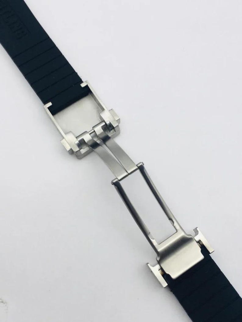 May include: A black rubber watch strap featuring a silver-toned stainless steel deployment clasp. The strap has a textured surface and the metal mechanism is shown in an open, extended position, revealing the hinge and locking components.