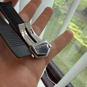 May include: A black rubber watch strap with a stainless steel clasp. The clasp is engraved with the brand name "TAG Heuer". The strap is held in a hand near a window with white blinds.