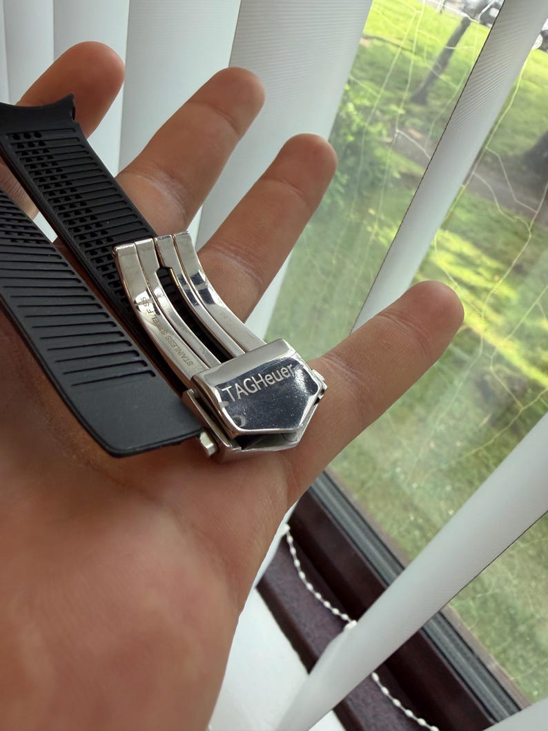 May include: A black rubber watch strap with a stainless steel clasp. The clasp is engraved with the brand name "TAG Heuer". The strap is held in a hand near a window with white blinds.
