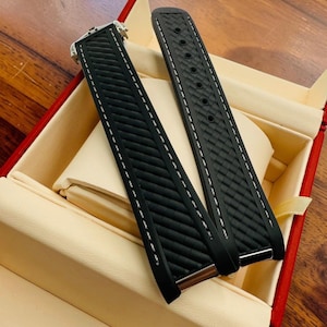 May include: Two black watch straps with white stitching and a textured pattern. The straps are displayed inside a red and beige box, suggesting a presentation or storage setting. The watch straps are likely for a high-end timepiece.
