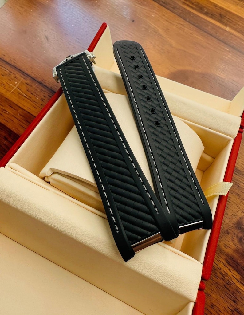 May include: Two black watch straps with white stitching and a textured pattern. The straps are displayed inside a red and beige box, suggesting a presentation or storage setting. The watch straps are likely for a high-end timepiece.