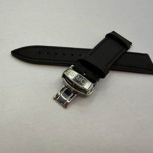 May include: A black watch strap featuring a silver-toned metal deployment clasp. The clasp is engraved with the letters ORS in a bold, sans-serif font. The strap is made of a smooth, matte material and includes a series of adjustment holes.