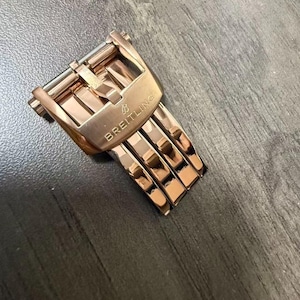 May include: A rose gold watch clasp with the brand name "BREITLING" engraved on the buckle. The clasp is rectangular with a series of interlocking links. The watch clasp is on a dark gray surface.