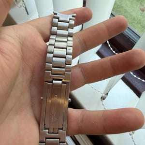 May include: A silver-toned metal watch band with a folding clasp. The band is composed of rectangular links and is held in a hand. The clasp is engraved with the brand name and other details.