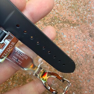 K&ouml;nnte beinhalten: Ein schwarzes Uhrenarmband mit einer silbernen Schlie&szlig;e. Die Schlie&szlig;e ist mit dem Markennamen "TUDOR" und dem Text "SWISS MADE STEELINOX EB" graviert. Das Armband besteht aus einem flexiblen Material und hat mehrere L&ouml;cher zur Anpassung.