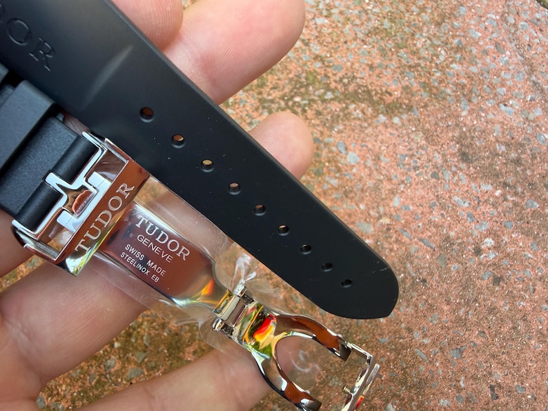 K&ouml;nnte beinhalten: Ein schwarzes Uhrenarmband mit einer silbernen Schlie&szlig;e. Die Schlie&szlig;e ist mit dem Markennamen "TUDOR" und dem Text "SWISS MADE STEELINOX EB" graviert. Das Armband besteht aus einem flexiblen Material und hat mehrere L&ouml;cher zur Anpassung.