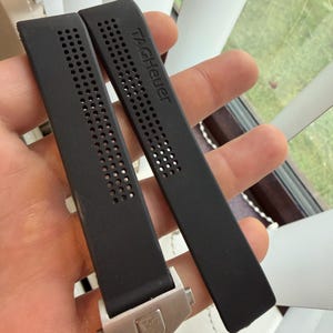 May include: Two black watch straps with the brand name "TAGHeuer" printed on one strap. The straps have a perforated design and a silver-colored clasp with the brand logo.