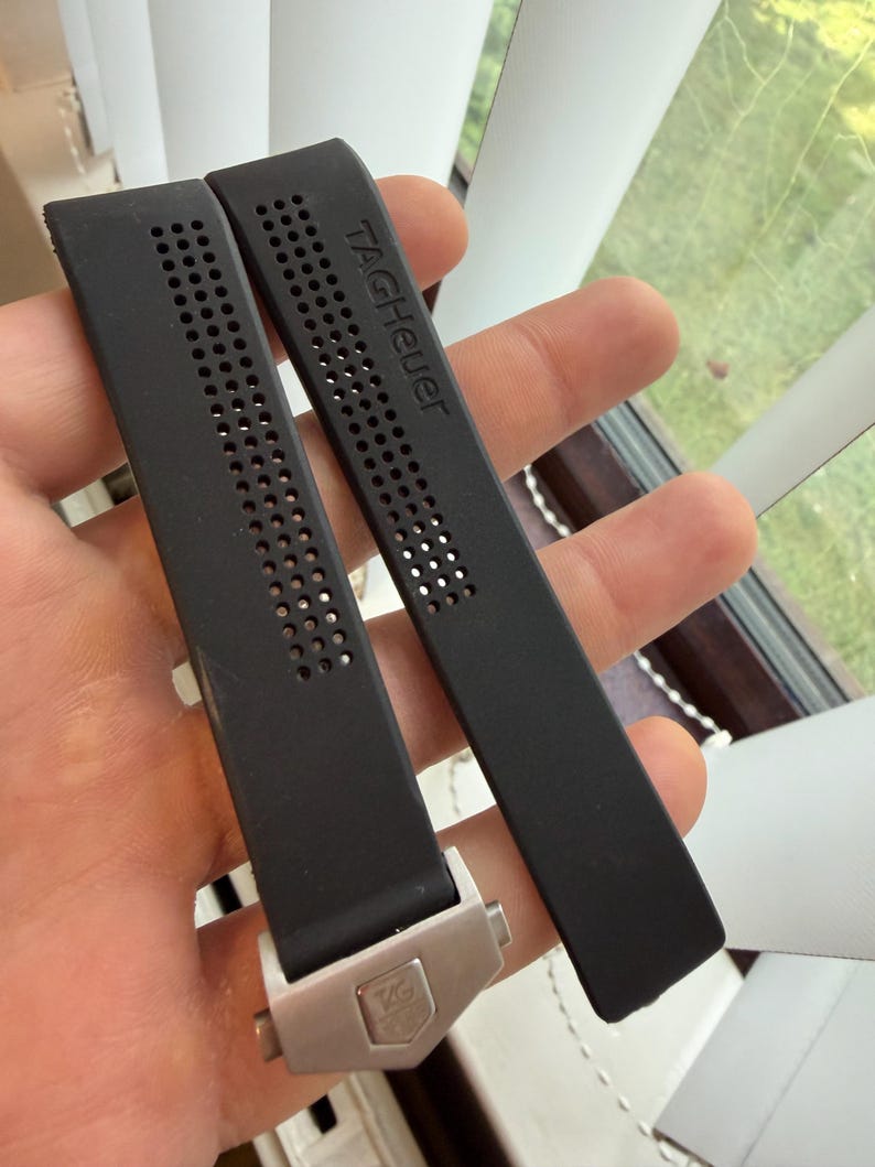 May include: Two black watch straps with the brand name "TAGHeuer" printed on one strap. The straps have a perforated design and a silver-colored clasp with the brand logo.