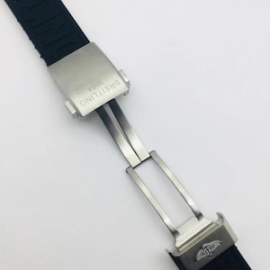 May include: A black rubber watch strap featuring a brushed stainless steel deployment clasp. The clasp is engraved with the text BREITLING 1884 and features the Breitling winged logo. The black strap has the brand name embossed along its length.