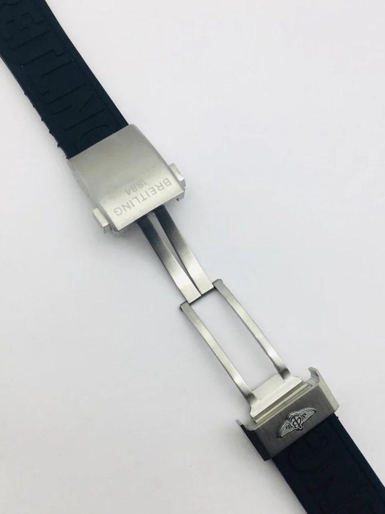 May include: A black rubber watch strap featuring a brushed stainless steel deployment clasp. The clasp is engraved with the text BREITLING 1884 and features the Breitling winged logo. The black strap has the brand name embossed along its length.