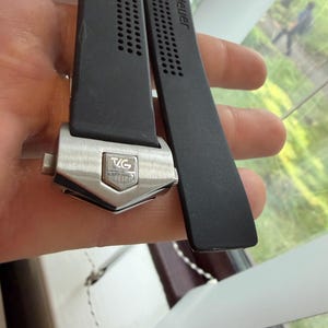 May include: Black watch strap with the brand name "TAG Heuer" printed in white. The strap has a silver-colored buckle with the brand's logo. The strap is made of a flexible material and has a series of small holes for ventilation.