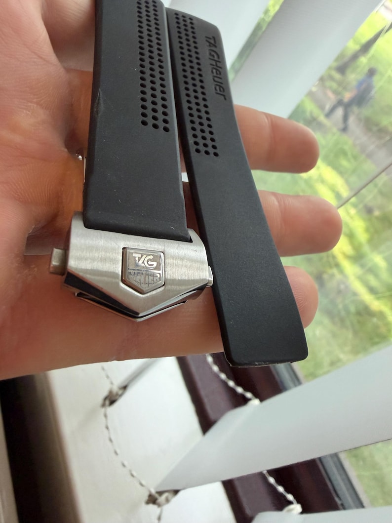 May include: Black watch strap with the brand name "TAG Heuer" printed in white. The strap has a silver-colored buckle with the brand's logo. The strap is made of a flexible material and has a series of small holes for ventilation.