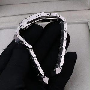 Pu&ograve; includere: Un cinturino per orologio in acciaio inossidabile color argento caratterizzato da una serie di maglie rettangolari interbloccate con un meccanismo di chiusura pieghevole sicuro. Il cinturino in metallo ha una finitura lucida e piccole viti sui lati.