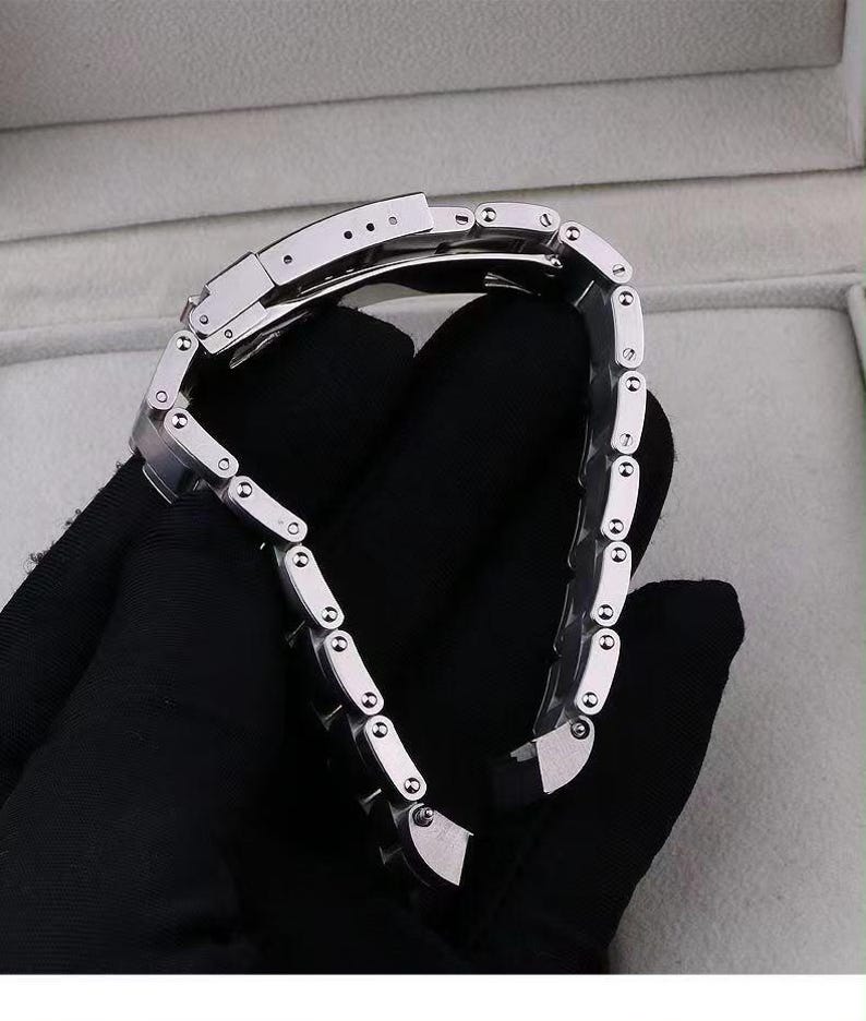 Pu&ograve; includere: Un cinturino per orologio in acciaio inossidabile color argento caratterizzato da una serie di maglie rettangolari interbloccate con un meccanismo di chiusura pieghevole sicuro. Il cinturino in metallo ha una finitura lucida e piccole viti sui lati.