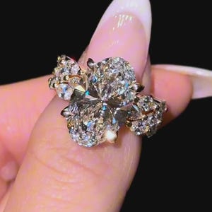 May include: A ring featuring a large, central oval-cut clear gemstone flanked by two smaller, pear-shaped clear gemstones on each side. The stones are set in a metallic band, creating a multi-stone design that catches light with a brilliant, reflective finish.