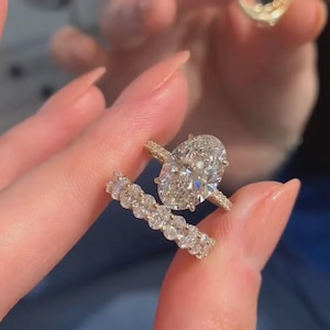 May include: A gold engagement ring featuring a large oval-cut centre stone with a pavé band, paired with a matching eternity band set with multiple round-cut stones. Both rings exhibit a high-shine metallic finish and clear, faceted gemstones.