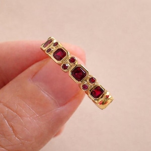 May include: A gold band ring featuring a series of square-cut and round-cut deep red rubies set along the outer surface. The metal has a polished finish, and the stones are arranged in a repeating pattern of larger square and smaller round gems.