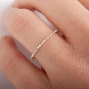 May include: A thin, delicate gold band ring featuring a continuous row of small, clear, round-cut pavé-set stones along the top half of the band. The metal has a warm, polished finish, and the stones are set closely together in a straight line.
