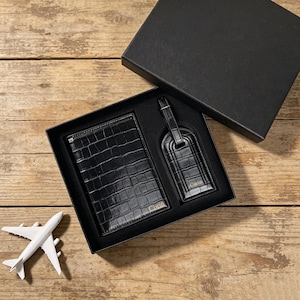 May include: A black passport holder and luggage tag set with a crocodile-embossed texture, both featuring the name Corin in gold lettering. The items are presented in a black gift box alongside a small white toy airplane model.