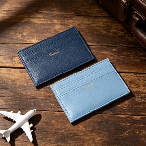 May include: Two rectangular card holders with a textured finish, one in dark navy blue with the gold embossed text Blond and one in light blue with the gold embossed text Seroa, accompanied by a small white toy airplane model.