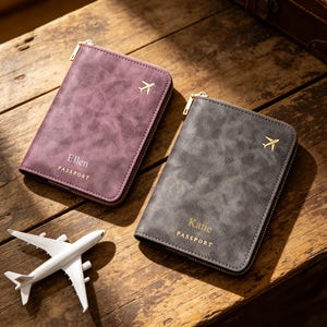 Könnte beinhalten: Zwei rechteckige Reisepasshüllen mit goldfarbenen Reißverschlüssen, eine in Pflaume und eine in Anthrazit. Jede zeigt ein goldenes Flugzeugsymbol, das Wort PASSPORT und einen in Gold geprägten Namen, Ellen oder Katie. Ein kleines weißes Spielzeugflugzeug.
