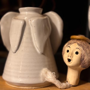 May include: A white ceramic angel-shaped oil lamp with a glossy finish and prominent wings. A separate ceramic head piece with a brown halo and painted facial features is connected to the base by a white fabric wick extending from the side.