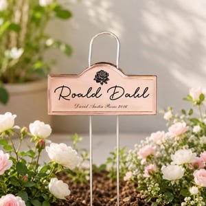 Puede incluir: Un marcador de jardín color cobre con el nombre "Roald Dahl" y "David Austin Roses 2016" en escritura negra. Un gráfico de rosa negra está encima del nombre. El marcador está en un jardín con rosas blancas y rosas.