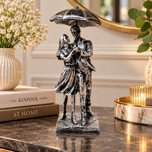 May include: A silver and black figurine depicting two figures standing together under a single umbrella. The sculpture features a textured finish, showing a person in a dress and a person in trousers, both positioned on a square base with a tiled pattern.