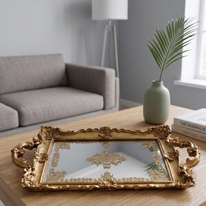 Op de afbeelding: Een rechthoekige, goudkleurige decoratieve dienblad met sierlijke details en handvatten. De dienblad heeft een spiegeloppervlak met een reliëf ontwerp. Het staat op een houten salontafel, met een vaas en boeken.