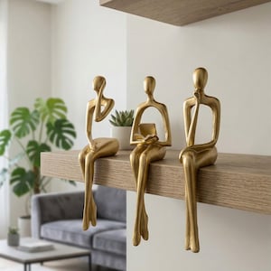 May include: Three gold-colored abstract human figurines in seated positions. The left figure rests a hand on its chin, the center figure holds an open book, and the right figure rests a hand on its chin. Each figure has elongated limbs and a smooth, metallic finish.