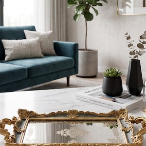 May include: A gold-framed mirrored tray with ornate handles sits on a marble coffee table. A teal sofa with two pillows is in the background, along with a potted plant, a black vase, and a succulent in a black pot. A book is also on the table.