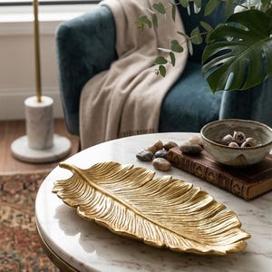 Könnte beinhalten: Eine goldfarbene, blattförmige Dekoschale liegt auf einem weißen Marmortisch. Die Schale hat detaillierte Adern und ist von einer kleinen Schale, einem Stapel Bücher und dekorativen Steinen umgeben. Ein türkisfarbener Sessel und eine beige Decke befinden sich im Hintergrund.