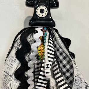 May include: A decorative tassel featuring a black ceramic telephone charm at the top, accented with white beads. The tassel consists of various fabric strips in black and white patterns, including gingham, stripes, and lace, with a small bead labeled H.